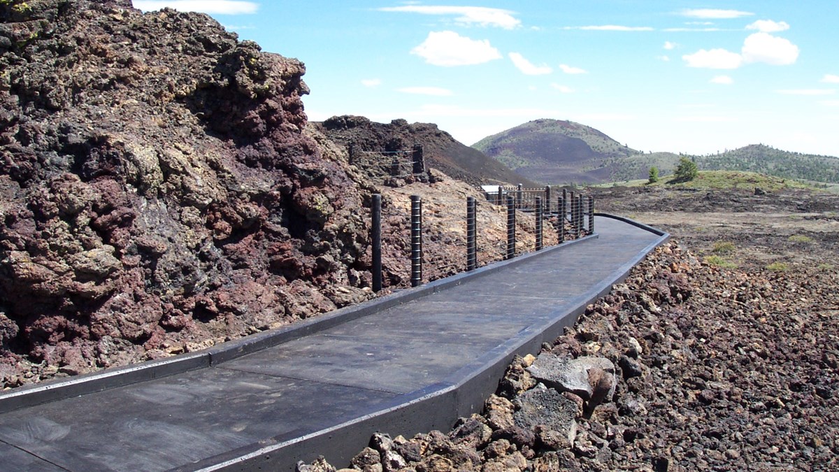 a black paved trail with some railing curving around a rocky outcrop with a large green and black cinder cone in the distance