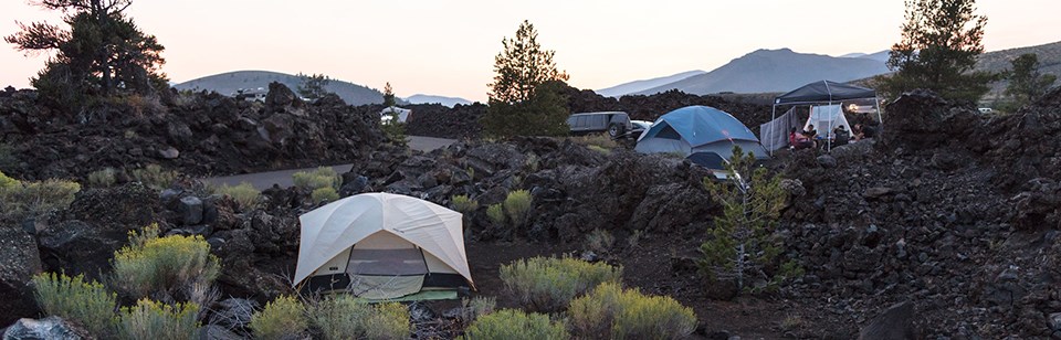 Tents are pitched among dark lava rocks, sagebrush, and a few pine trees