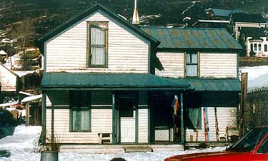 This small, 1 1/2 story house in a hilly historic mining community in the West was built in 1891. The wood frame building with an L-plan features a prominent gable roof, a one-story porch across the facade, and two historic sheds at the rear. Photos: NPS files.