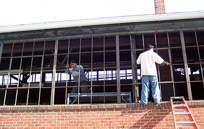 Two men repairing steel multi-light windows on a brick industrial building.