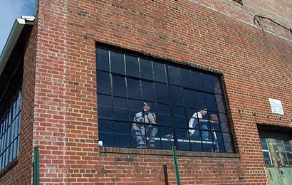 Two men repairing steel multi-light windows from the inside of a brick industrial building.