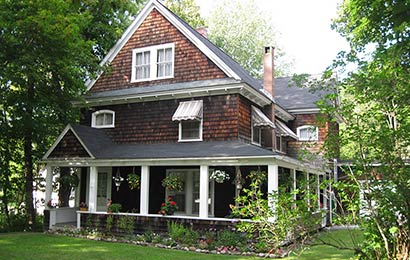 House with brown shingle siding, a wrap-around porch, and awnings over the second floor windows.