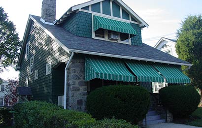 Bungalow with green and white striped awnings between stone porch pillars and on the second floor dormer window.