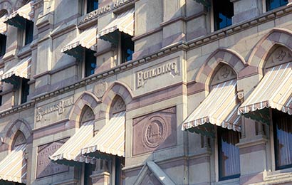 Portion of the façade of a brown and beige stone building with brown and beige awnings at each window.