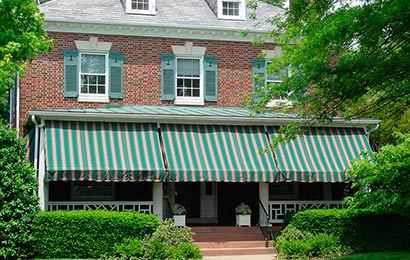 Red brick house with two dormers in a slate roof, green shutters and a green and white striped awning on its porch.