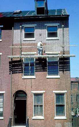 This is an image of a brick building being insensitively repointed. The visual character of the front wall is being changed from a wall where the bricks predominate to a wall that is visually dominated by the mortar joints. The worker is apparently unaware of the problem. Photo: NPS files.