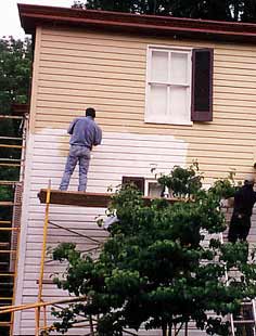 This is an image of a worker on scaffolding priming the exterior of the wood siding on a historic house prior to applying a finish coat. One shutter has been removed for work as well. Photo: NPS files.