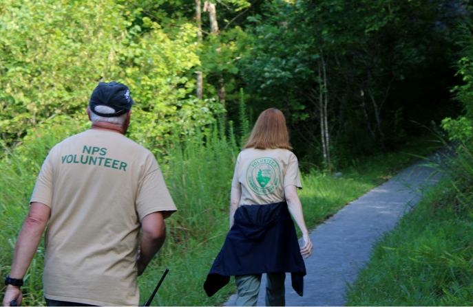 back view of a man and woman wearing NPS volunteer shirts hiking up a paved pathway into the woods