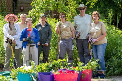 Seven volunteers pose in front of a garden bed with piles of weeds they have pulled