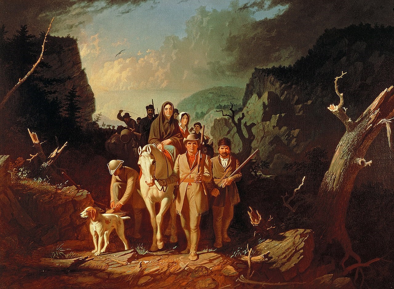 A group of people and animals being lead by a man in a hat through a gap in the mountain.