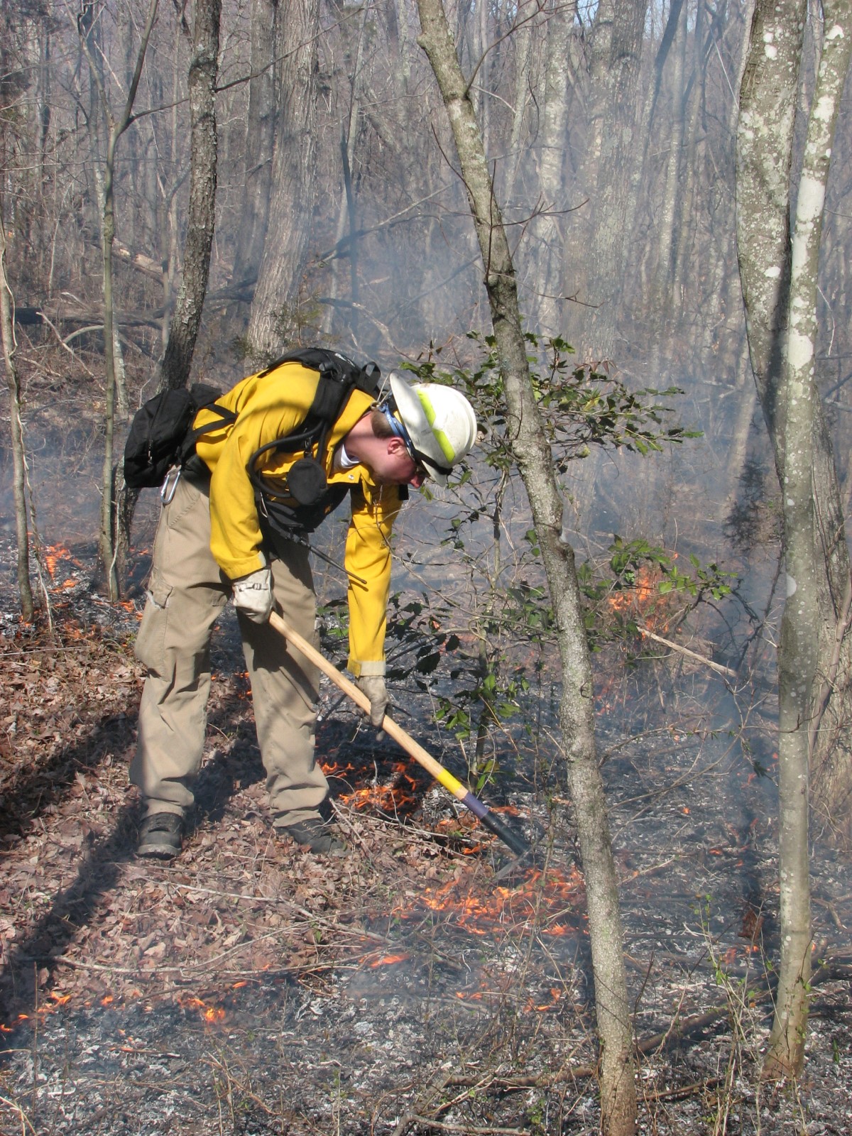 A Wildland Firefighter works to control a prescribed burn at Cumberland Gap National Historical Park A man wearing protective gear scrapes burning leaves in the forest with a pickaxe