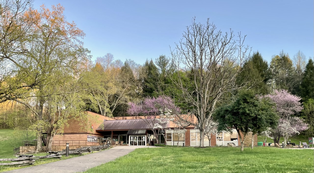 A sidewalk along a split rail fence leads up to a large building surrounded by trees.