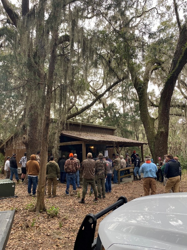 Group of hunters stand facing a wooden structure with a porch on it under large trees