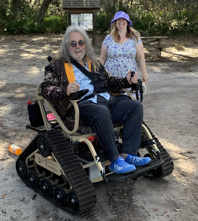 a woman in a blue flowered dress stands behind a man in a motorized track chair