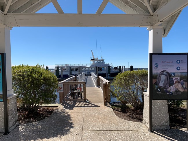 Bushes on either side of a white gazebo frame an open gate to a boardwalk leading to a passenger ferry