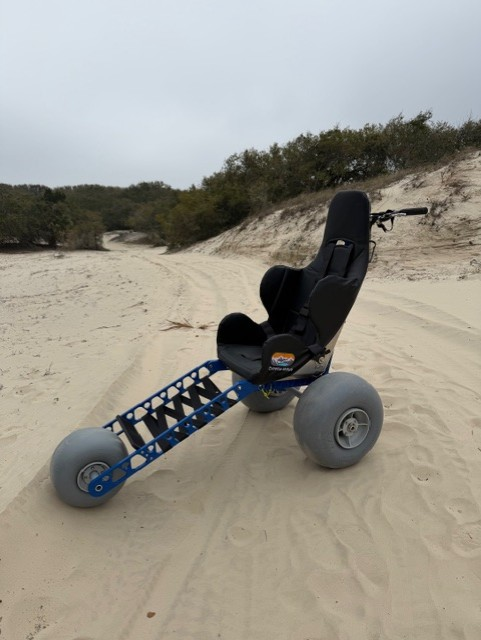 A three-wheeled balloon tire non-motorized wheel chair sitting on a sandy road with sand dunes covered in plants in the background.
