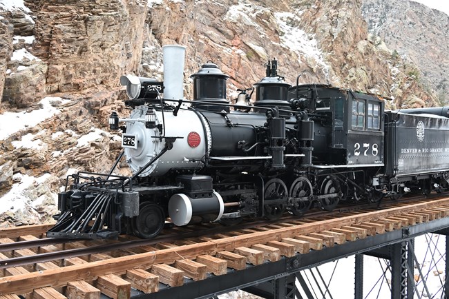 A black and gray steam engine train car sits on a wooden platform. Other train cars are behind it. Snow and canyon walls are in the background.