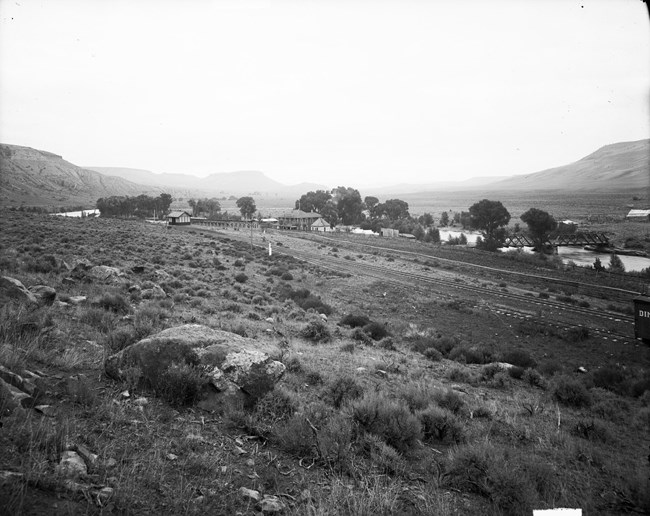 Historic black and white image of buildings, railroad tracks, a river, bridges, and mesas in the background