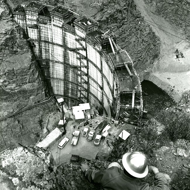 Historic image of a cosntruction worker sitting on a ledge above a partially constructed dam. The dam is multiple stories high with scaffolding and equipment on it. Trucks are near the bottom. Canyon walls surround it.