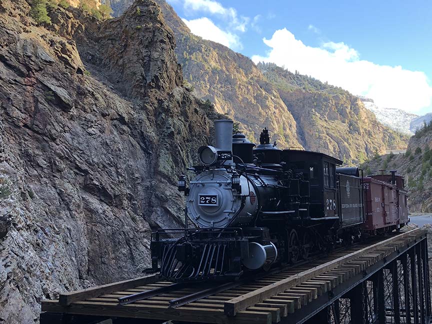 Narrow gauge engine, tender, boxcar and caboose sit on a railroad bridge in a deep, dark canyon over a rushing stream