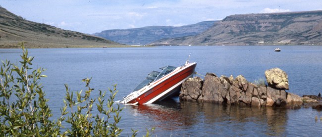 A damaged motorboat leans up against rocks rising out of a lake.