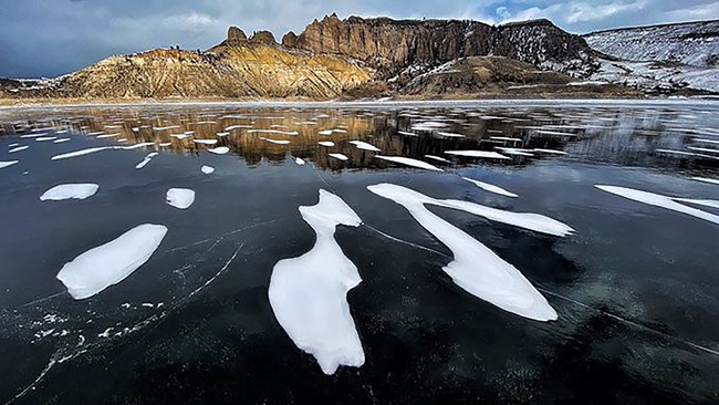 Patches of snow upon the frozen surface of a lake with craggy peaks in the background