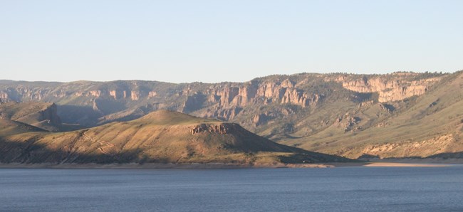 A hilly and rocky landscape of green and brown has a blue body of water in the foreground and clear blue skies in the background