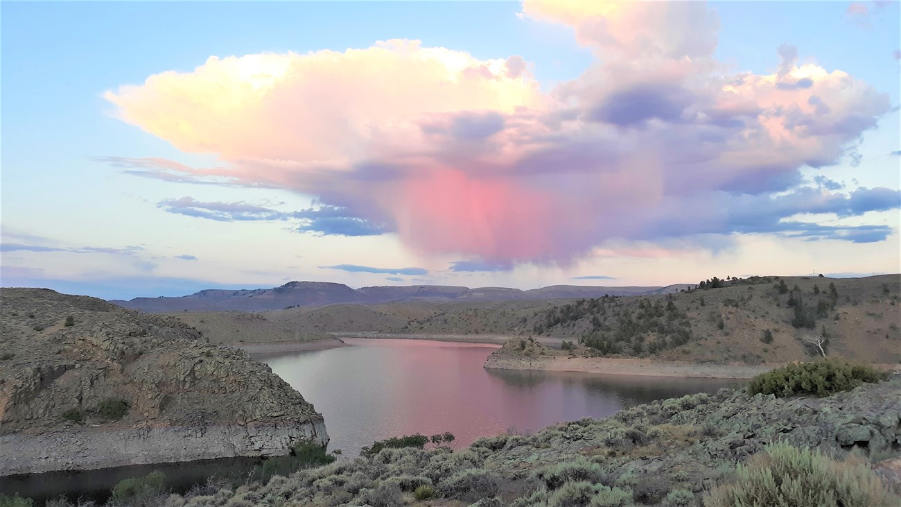 A pink cotton candy colored clouds floats above a glassy surface of still water the size of the large lake. The color reflects in the surface. The landscape surrounding the water is dotted in sage colored bushes.