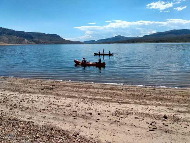 Two groups of people paddling canoes on a large reservoir with a rocky beach in the foreground