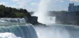blueish-green water crashes over a fall creating white foam & mist, a cityscape fades in the background