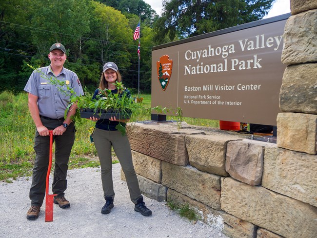 A male uniformed ranger and a woman in a different uniform stand smiling next to a sign for Cuyahoga Valley National Park; the ranger holds an orange planting tool and the woman holds a tray of green plants.
