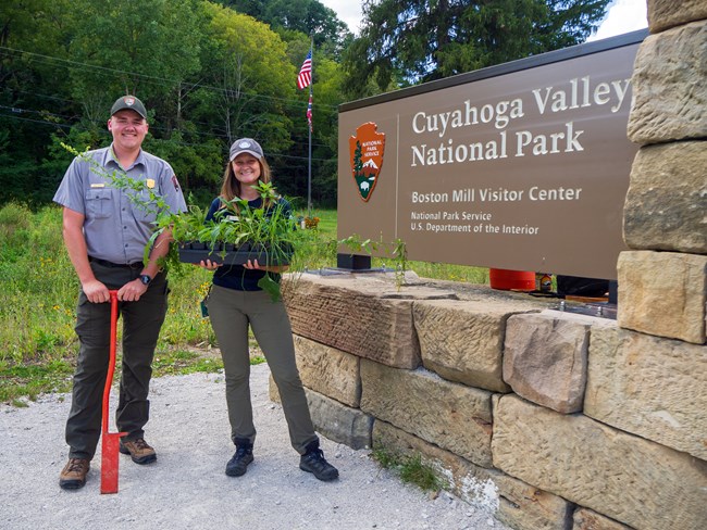 A male uniformed ranger and a woman in a different uniform stand smiling next to a sign for Cuyahoga Valley National Park; the ranger holds an orange planting tool and the woman holds a tray of green plants.