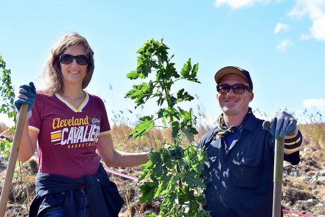 Two people smile on a sunny day, standing on either side of a small tree with green leaves.