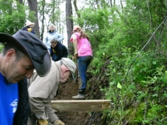 Six people stand along a dirt path through green plants, using hand tools.