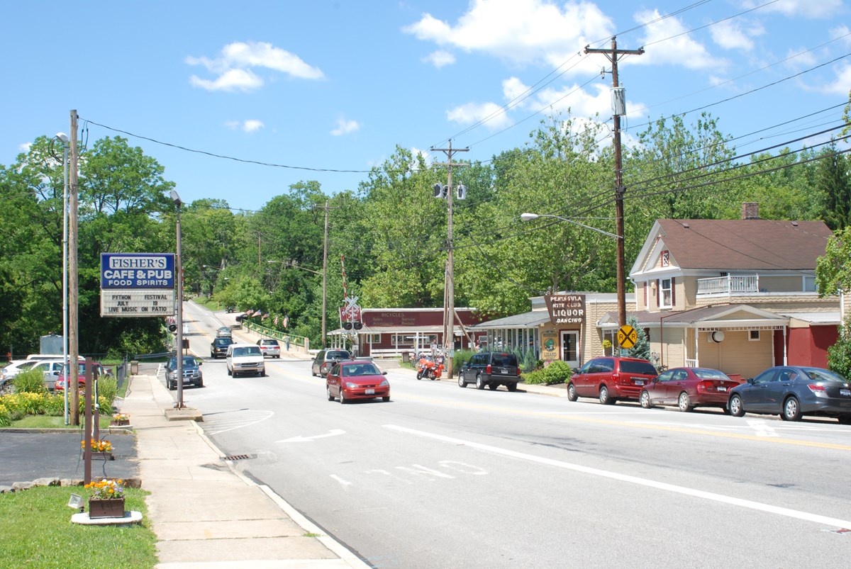 Vehicles drive down a busy two lane road, crossing a bridge. Others are parked along the sides. Signs advertise restaurants and other businesses in historic wood buildings with trees in the background.