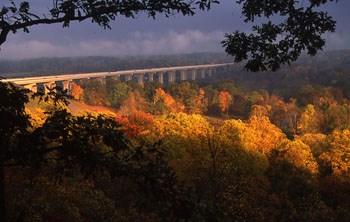 Orang and yellow autumn leaves illuminated in a river valley spanned by a gray concrete bridge.