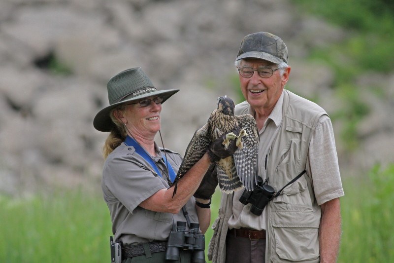 A female uniformed ranger holds a falcon in her gloved hands; next to her a man in khaki vest smiles and looks at the bird.