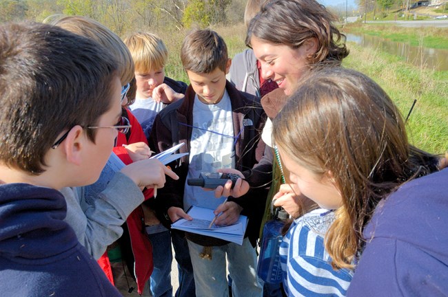A group of students and an adult, some holding paper and pencil, stand in a circle looking down at a handheld electronic device.