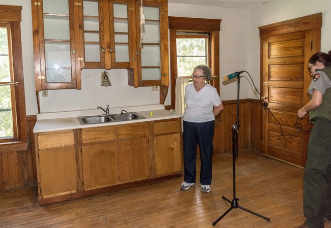 A woman stands in front of a microphone in a kitchen with wooden cabinets