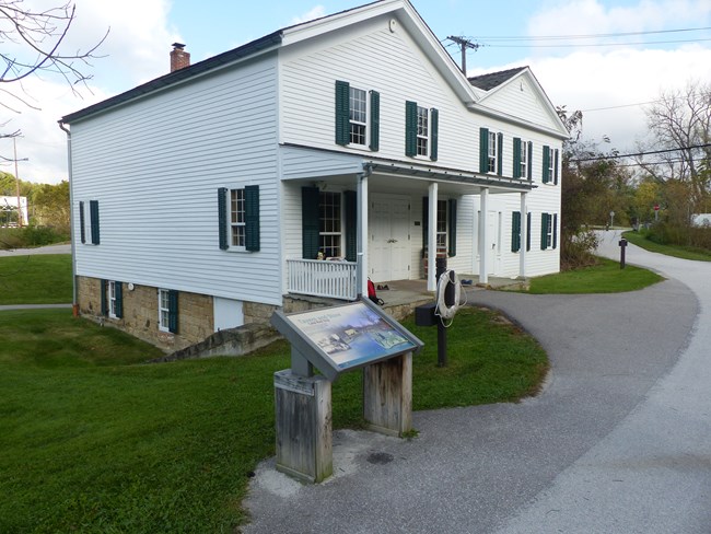 A graphic panel and life ring stand outside a two story white building with green shutters, a porch and an M-shaped roofline.