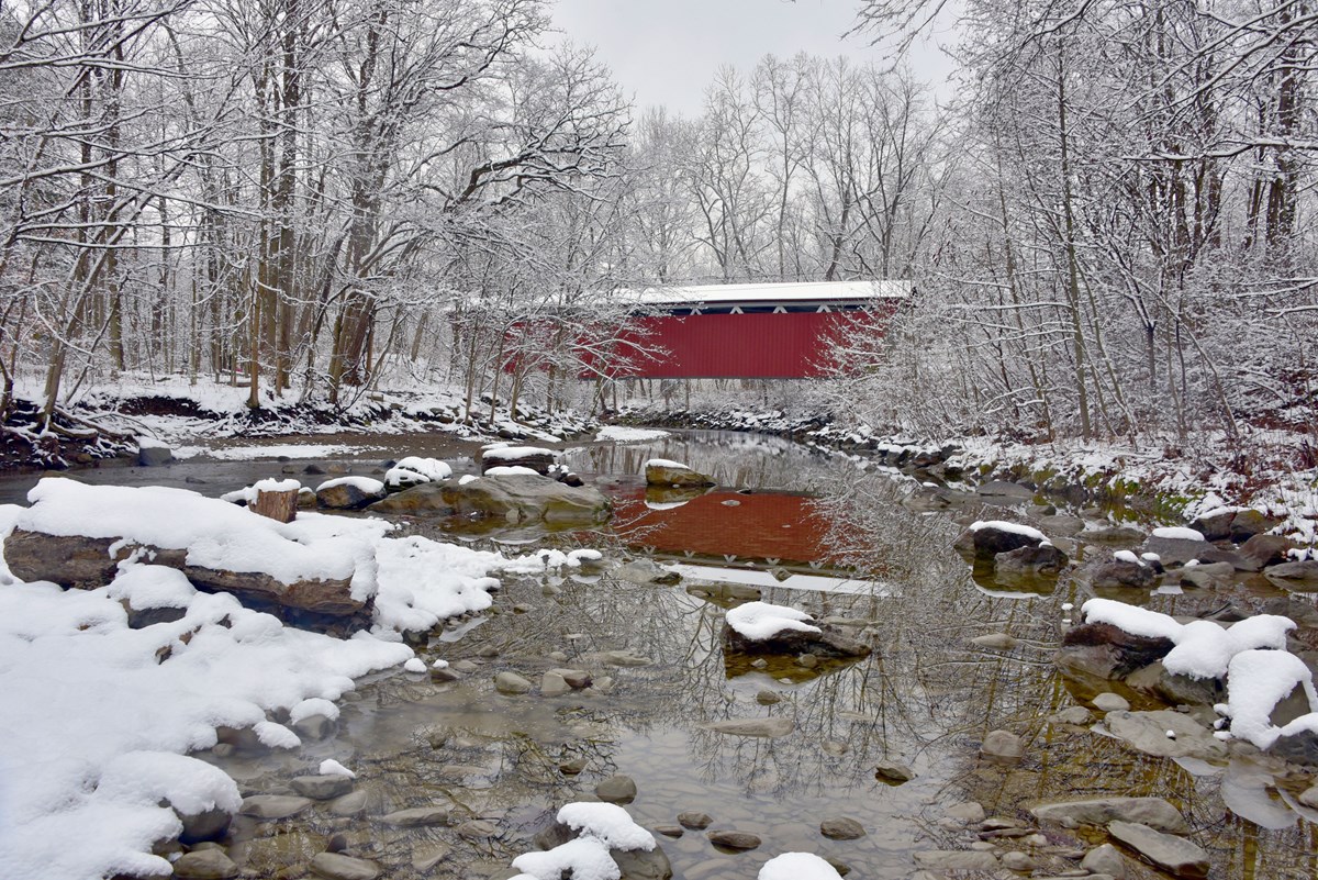 A snowy scene of a rocky stream flowing under a red covered bridge with woods on either bank.
