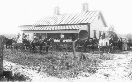 Black and white photo of Sumner Creamery, in 1902. A family and farmers, nine people total, pose in front of a barn-style home with a large porch and an awning. There are four wagons full of milk, the each wagon drawn by two large, brown horses.