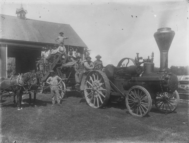 Black and white photo of about dozen men and boys standing and sitting on a mechanical wagon with a large chimney, in front of an open barn.