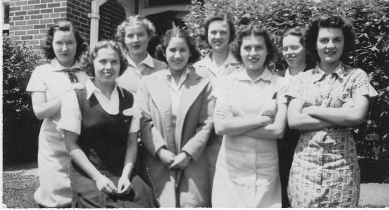 Historic photo of eight young women students standing in front of a brick building.