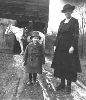 Historical photo a woman and child dressing in nice clothing standing on a muddy road with a man on a horse and a barn in the background.