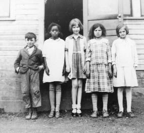 Black and white photo of Everett schoolhouse and five students. One is a boy, standing on the far left. The other four are girls, all in dresses. There are 4 white children and one black girl standing next to the boy. All look about 8 years old.