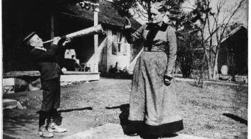 Black and white photo of a young boy and middle-aged woman in long dress; the boy holds a long, conical instrument to his mouth and points it toward the woman, who looks sternly at the boy and holds up her hand.