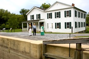Three volunteers wearing traditional 1800's clothing stand besides the canal lock mechanism in front of the historic Canal Exploration Center building.