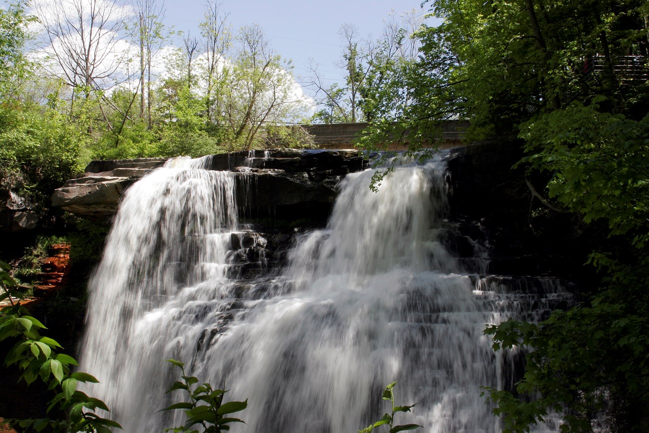 Long exposure photo shows white water cascading over the top of a waterfall; green tree branches and blue sky frame the image.