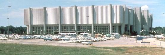 A large, square building surrounded by a parking lot full of 1970s-era cars and buses.